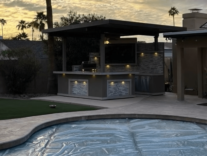 The Kokomo Grills Best Outdoor Kitchen Bermuda BBQ Island with TV sits under a pavilion roof beside a pool with a blue tarp, accented by stylish lights. Palm trees and a house frame the sunset in the background.