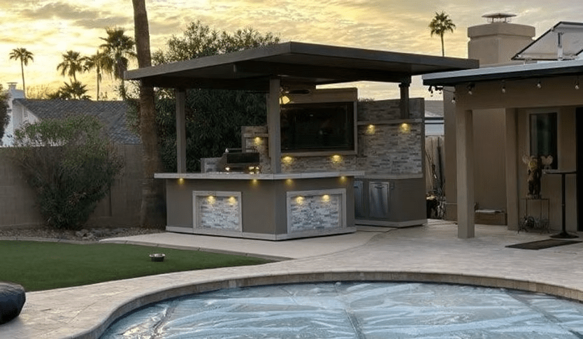 A backyard showcases a covered outdoor kitchen and bar with modern stone accents, a Kokomo Grills Best Outdoor Kitchen Bermuda BBQ Island with TV, under-counter lighting, a pool with cover, all surrounded by patio and landscaping at sunset.