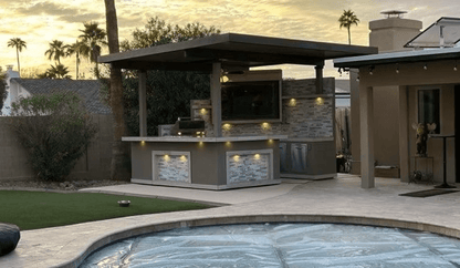 A backyard showcases a covered outdoor kitchen and bar with modern stone accents, a Kokomo Grills Best Outdoor Kitchen Bermuda BBQ Island with TV, under-counter lighting, a pool with cover, all surrounded by patio and landscaping at sunset.