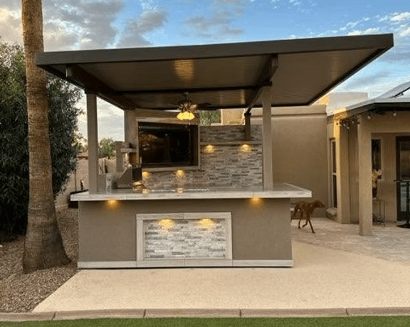 The Kokomo Grills Best Outdoor Kitchen Bermuda BBQ Island with TV features a roof, stone bar counter, ceiling fan, and built-in lights—ideal for backyards with pets and palm trees.
