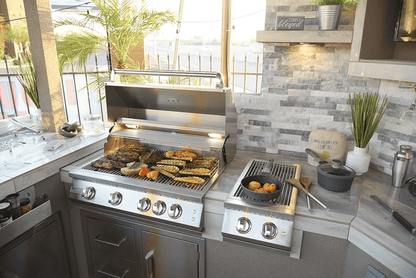 Outdoor kitchen with a Kokomo Grills Best Outdoor Kitchen Bermuda BBQ Island with TV, stainless steel grill cooking meats and veggies, side burner for pots and pans, plus potted plants and decor on a stone countertop and backsplash.