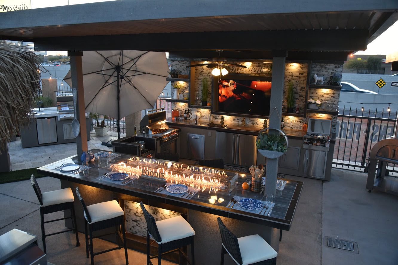 Experience dusk dining with the Kokomo Grills Best Outdoor Kitchen Bermuda BBQ Island with TV—featuring bar seating, a long counter fire feature, umbrella, ceiling fan, appliances, and warm lighting under a pergola.