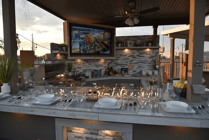 A stylish outdoor kitchen featuring the Kokomo Grills Best Outdoor Kitchen Bermuda BBQ Island with TV, marble countertop, stainless steel appliances, place settings, gas fire feature, and decorative plants under a covered patio.