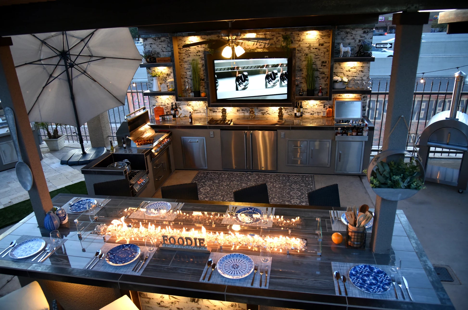 The Kokomo Grills Best Outdoor Kitchen Bermuda BBQ Island with TV features stainless steel appliances, a grill, sink, bar, dining table with blue and white plates, and a fire feature; decorated shelves and lighting add cozy modern style.