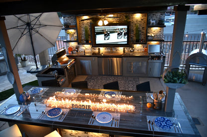 The Kokomo Grills Best Outdoor Kitchen Bermuda BBQ Island with TV features stainless steel appliances, a grill, sink, bar, dining table with blue and white plates, and a fire feature; decorated shelves and lighting add cozy modern style.
