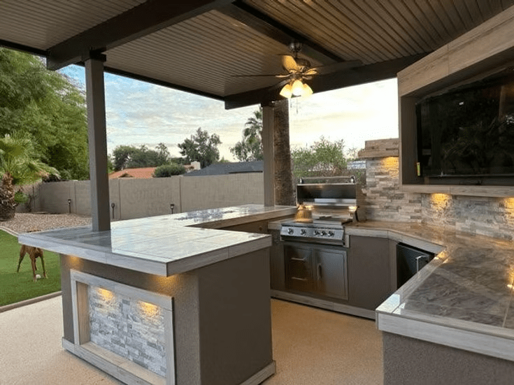 Modern outdoor kitchen with the Kokomo Grills Best Outdoor Kitchen Bermuda BBQ Island with TV, featuring a stone countertop, built-in grill, ceiling fan, under-cabinet lighting, and inviting views of trees and a brick wall under a covered patio.