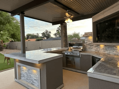 Modern outdoor kitchen with the Kokomo Grills Best Outdoor Kitchen Bermuda BBQ Island with TV, featuring a stone countertop, built-in grill, ceiling fan, under-cabinet lighting, and inviting views of trees and a brick wall under a covered patio.