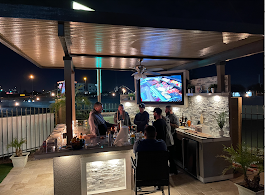 At night, people gather around the Kokomo Grills Best Outdoor Kitchen Bermuda BBQ Island with TV by Kokomo Grills. The well-lit area features barstools, drinks, plants, and city lights shining in the background.