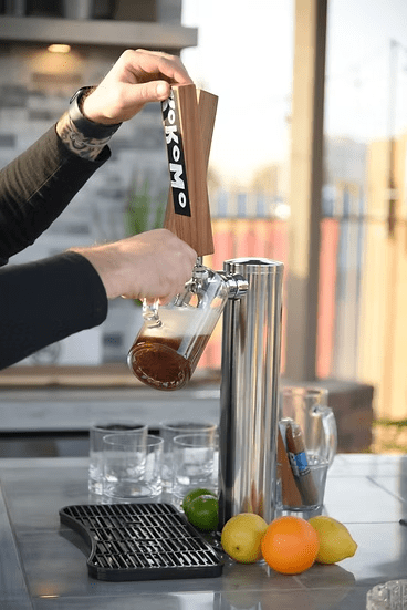 A person pours draft beer from a tap into a glass mug, holding a Kokomo Grills can. On the counter—lemons, lime, cigar, and empty glasses hint at gatherings by the Kokomo Grills Best Outdoor Kitchen Bermuda BBQ Island with TV.