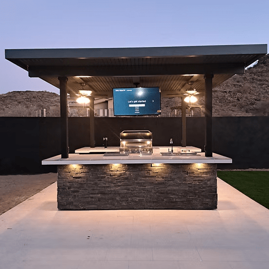 The Kokomo Grills Big Island Sports Bar Outdoor Kitchen Grill by Kokomo Grills features a stone countertop, built-in BBQ island, overhead lights, and TV under a sleek black roof—ideal for a backyard sports bar vibe in your desert landscape.