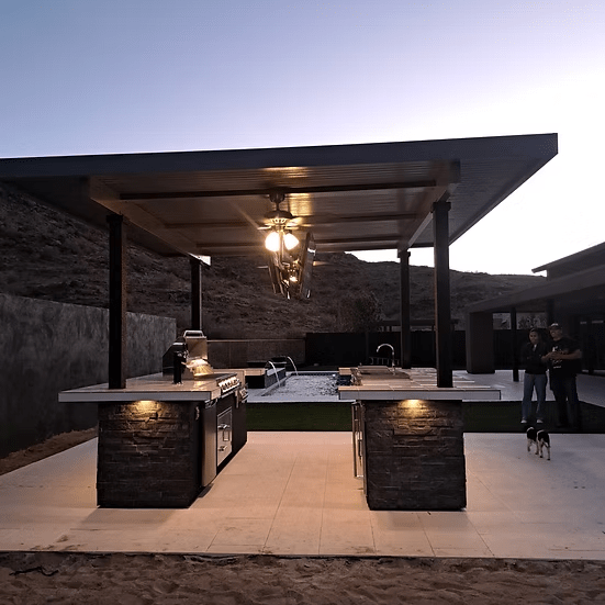 The Kokomo Grills Big Island Sports Bar Outdoor Kitchen Grill by Kokomo Grills, featuring a metal roof, island, countertops, and lighting, stands on the patio near the pool at dusk as two people and a dog enjoy the hilltop view.