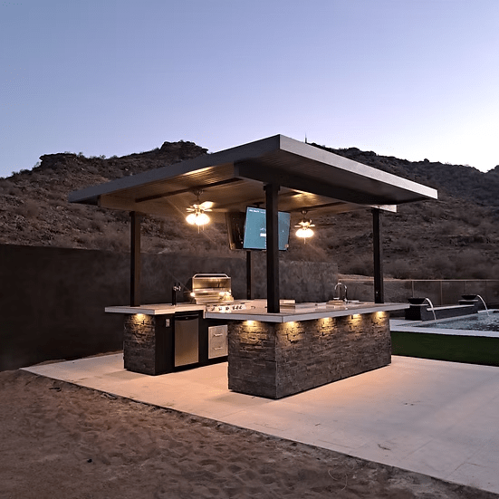 Enjoy the Kokomo Grills Big Island Sports Bar Outdoor Kitchen Grill by Kokomo Grills featuring a stone BBQ island, built-in grill, sink, bar seating, fans, TV, and roof—your perfect desert oasis for entertaining at sunset.