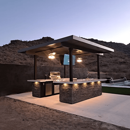 Enjoy the Kokomo Grills Big Island Sports Bar Outdoor Kitchen Grill by Kokomo Grills featuring a stone BBQ island, built-in grill, sink, bar seating, fans, TV, and roof—your perfect desert oasis for entertaining at sunset.