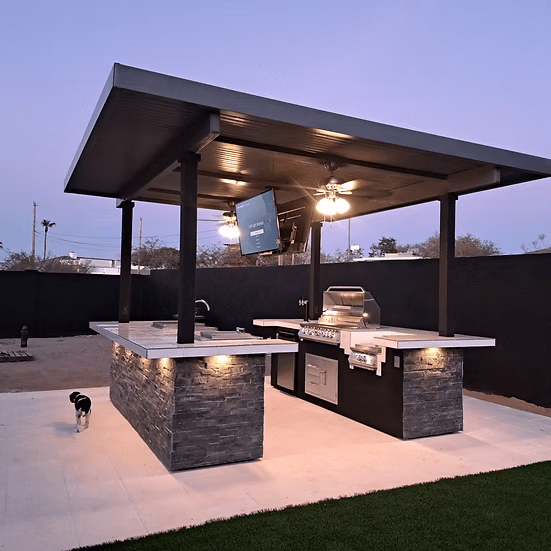 A modern outdoor kitchen with stone counters and stainless steel appliances under a covered pergola features the Kokomo Grills Big Island Sports Bar Outdoor Kitchen Grill. Ceiling fans and lights brighten the space, where a black and white dog stands at dusk.