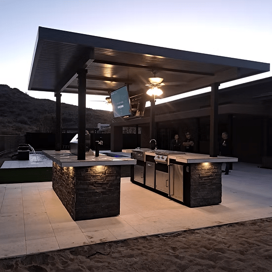 A modern outdoor kitchen features stone counters and the Kokomo Grills Big Island Sports Bar Outdoor Kitchen Grill under a covered patio at dusk, with three people in the background and mountains rising behind the scene.