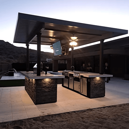 A modern outdoor kitchen features stone counters and the Kokomo Grills Big Island Sports Bar Outdoor Kitchen Grill under a covered patio at dusk, with three people in the background and mountains rising behind the scene.