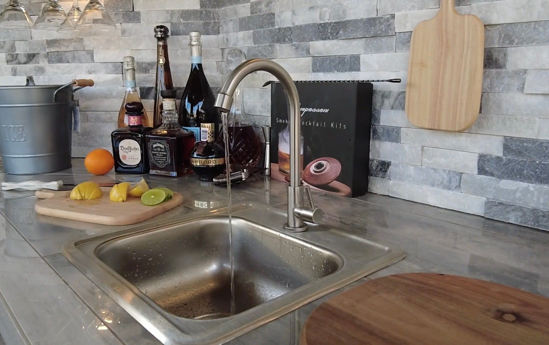 A modern kitchen counter features the Kokomo Grills Built-In 15 x 15 Outdoor Kitchen Sink by Kokomo Grills, cocktail ingredients like lemon and lime slices on cutting boards, bottles of alcohol, a cocktail kit, and a gray tile backsplash.