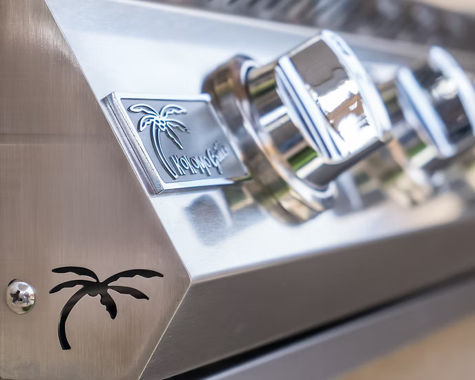 Close-up of a shiny Kokomo Grills Built-In Double Side Burner in stainless steel with chrome knobs, engraved palm tree logo, removable cover, and a palm tree cutout on the corner panel.