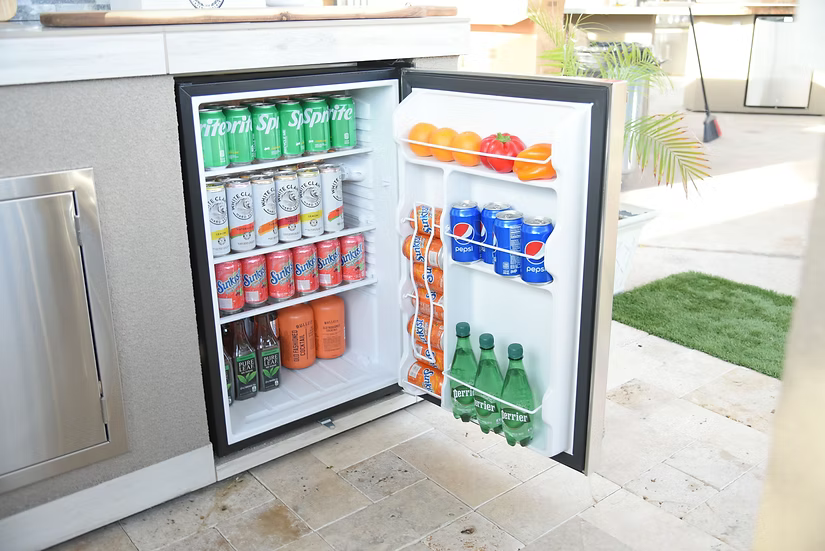A Kokomo Grills Built-In Outdoor Kitchen Refrigerator with Temp Control, Soda Rack, and Lights (KO-FRIDGE) sits on a tiled patio near greenery, stocked with Sprite, San Pellegrino, fruit, and eggs on the door shelves.