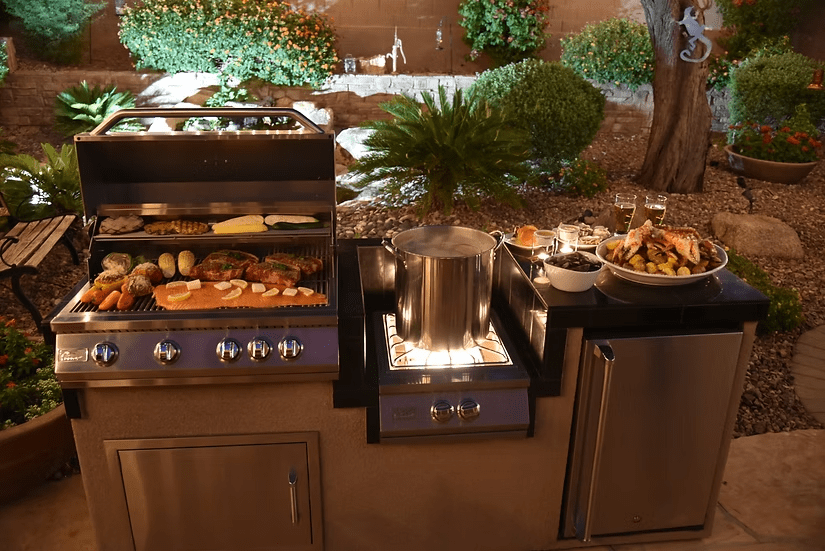 A Kokomo Grills Built-In Power Burner with a removable grate for wok, plus a stainless steel grill cooking meats and veggies, stands in a landscaped backyard at night with a large pot boiling and grilled food on a platter.