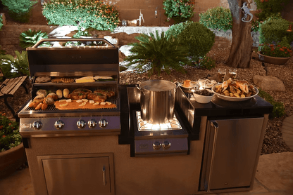 A Kokomo Grills Built-In Power Burner with a removable grate for wok, plus a stainless steel grill cooking meats and veggies, stands in a landscaped backyard at night with a large pot boiling and grilled food on a platter.