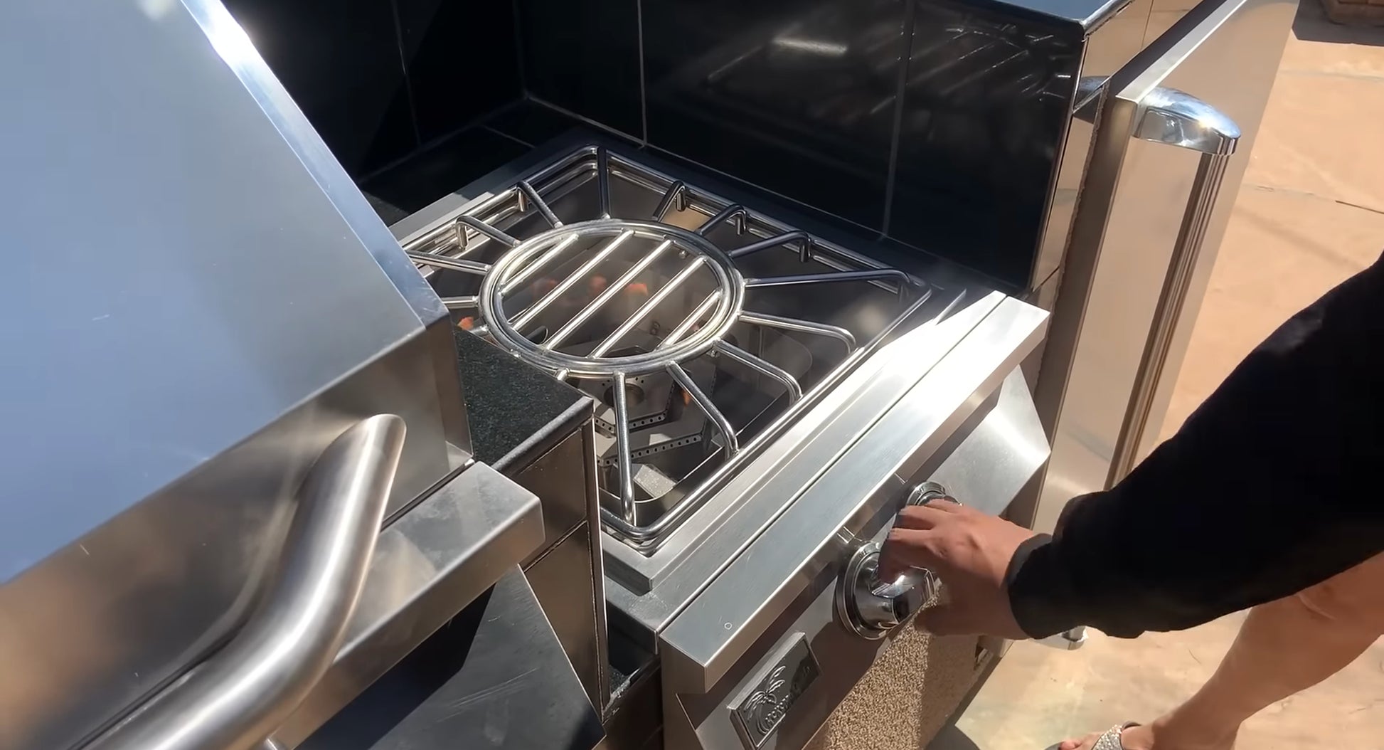 A person turns the knob on a Kokomo Grills Built-In Power Burner with Removable Grate for Wok in their outdoor kitchen, ready to ignite it. Only their arm and part of their legs are visible.