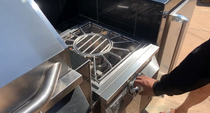 A person turns the knob on a Kokomo Grills Built-In Power Burner with Removable Grate for Wok in their outdoor kitchen, ready to ignite it. Only their arm and part of their legs are visible.