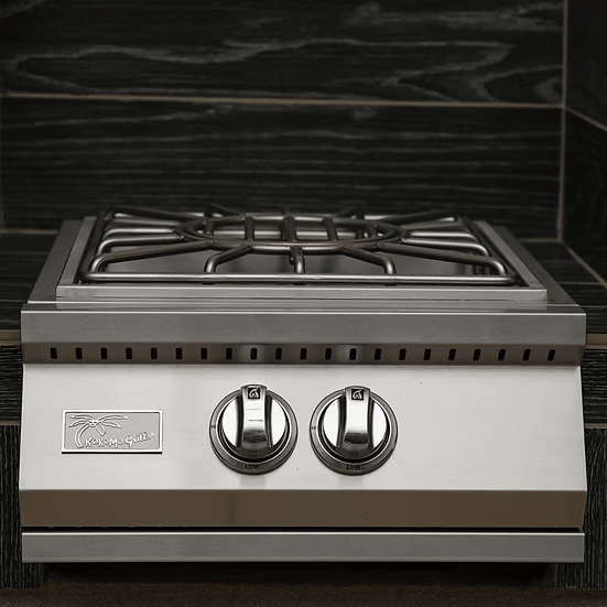 The Kokomo Grills Built-In Power Burner with Removable Grate for Wok is a stainless steel gas stove featuring two burners, each operated by a large round front knob, and set against a dark wood-patterned backsplash.