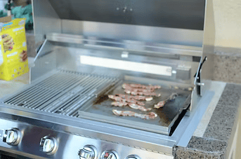 A Kokomo Grills 5 Burner 40-Inch Cart Model BBQ Grill with Locking Casters sizzles bacon on the griddle plate, while a box of frozen waffles sits on the countertop nearby.