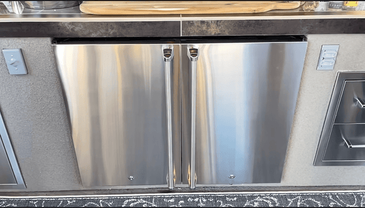 The Kokomo Grills Double Door Outdoor Refrigerator DBLFRDGE features two stainless steel doors with vertical handles, set into a kitchen counter. A patterned rug is visible on the floor below.