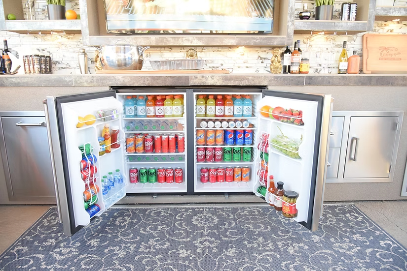 Two Kokomo Grills DBLFRDGE Double Door Outdoor Refrigerators by Kokomo Grills, doors open, display organized drinks, fruits, and condiments in a modern outdoor kitchen. Canned sodas, sports drinks, and water bottles are arranged above a patterned rug.