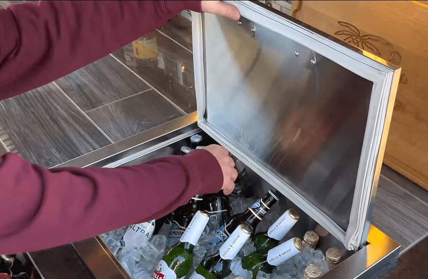 A person in a red long-sleeve shirt opens the Kokomo Grills Drop-In Stainless Steel Ice Chest 23 x 17 KO-AIC, filled with ice and assorted bottled drinks, on an indoor tiled surface.