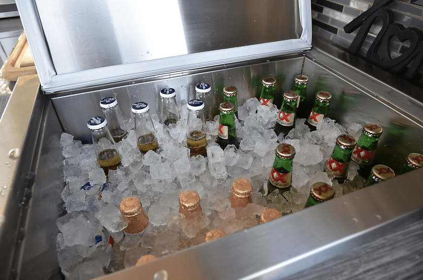 The Kokomo Grills Drop-In Stainless Steel Ice Chest 23 x 17 KO-AIC by Kokomo Grills is open, filled with ice and assorted bottled beers—some with blue or green caps, and others featuring gold foil tops—all visible inside.
