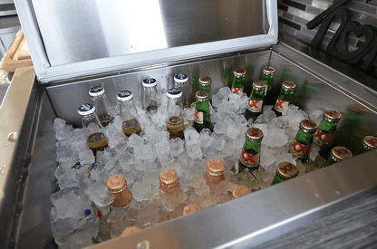 The Kokomo Grills Drop-In Stainless Steel Ice Chest 23 x 17 KO-AIC by Kokomo Grills is open, filled with ice and assorted bottled beers—some with blue or green caps, and others featuring gold foil tops—all visible inside.