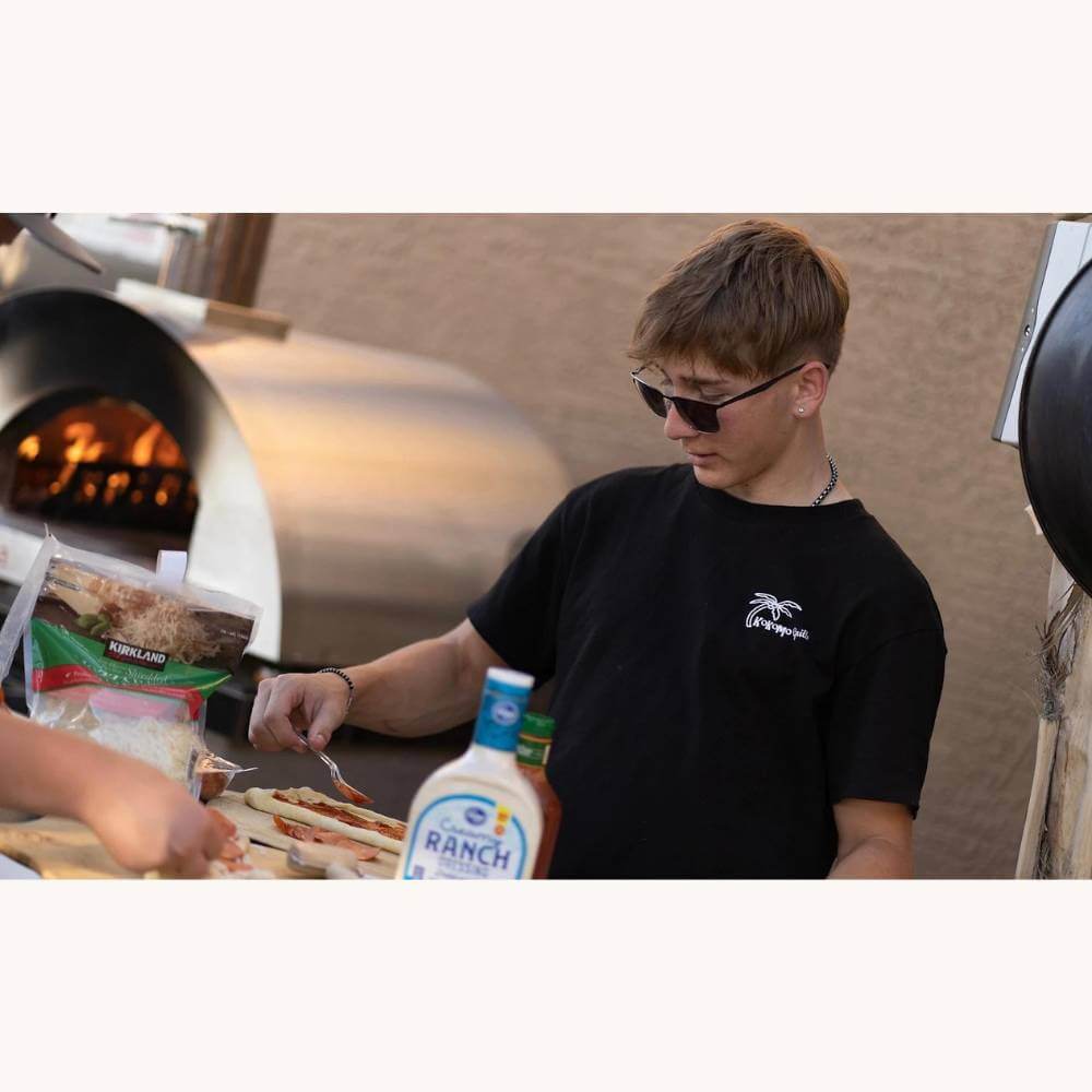 Young man wearing sunglasses making pizza outdoors, with the Kokomo 32” Dual Fuel Freestanding Pizza Oven fired up in the background.