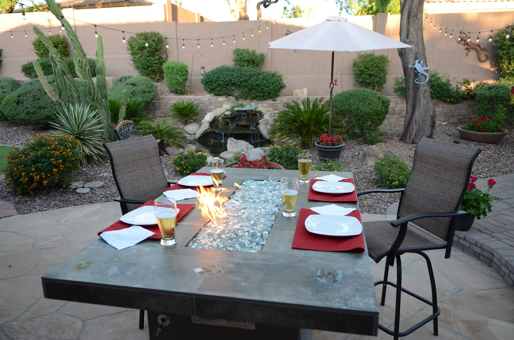 A Kokomo Grills Entertainer Bar Gas Fire Pit Table with Fire-Glass is surrounded by a patio dining set for four with drinks and red napkins. The backyard features greenery, a waterfall, string lights, and an umbrella shading one chair.