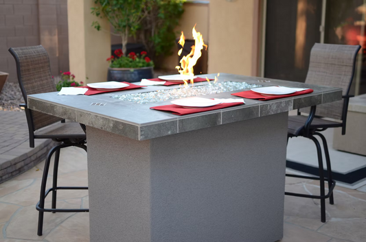 The Kokomo Grills Entertainer Bar Gas Fire Pit Table with Fire-Glass Entertainer-FP is surrounded by four brown chairs, set with red placemats, white plates, and napkins, with patio and plants in the background.