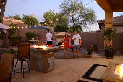At sunset, four adults relax around the Kokomo Grills Entertainer Bar Gas Fire Pit Table with Fire-Glass. String lights glow overhead while fireglass sparkles in the inviting firepit, creating a warm and welcoming backyard atmosphere.