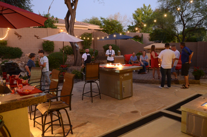 A group gathers at dusk on a well-lit patio, chatting around the Kokomo Grills Entertainer Bar Gas Fire Pit Table with Fire-Glass by Kokomo Grills. String lights and umbrellas decorate the charming, landscaped space.