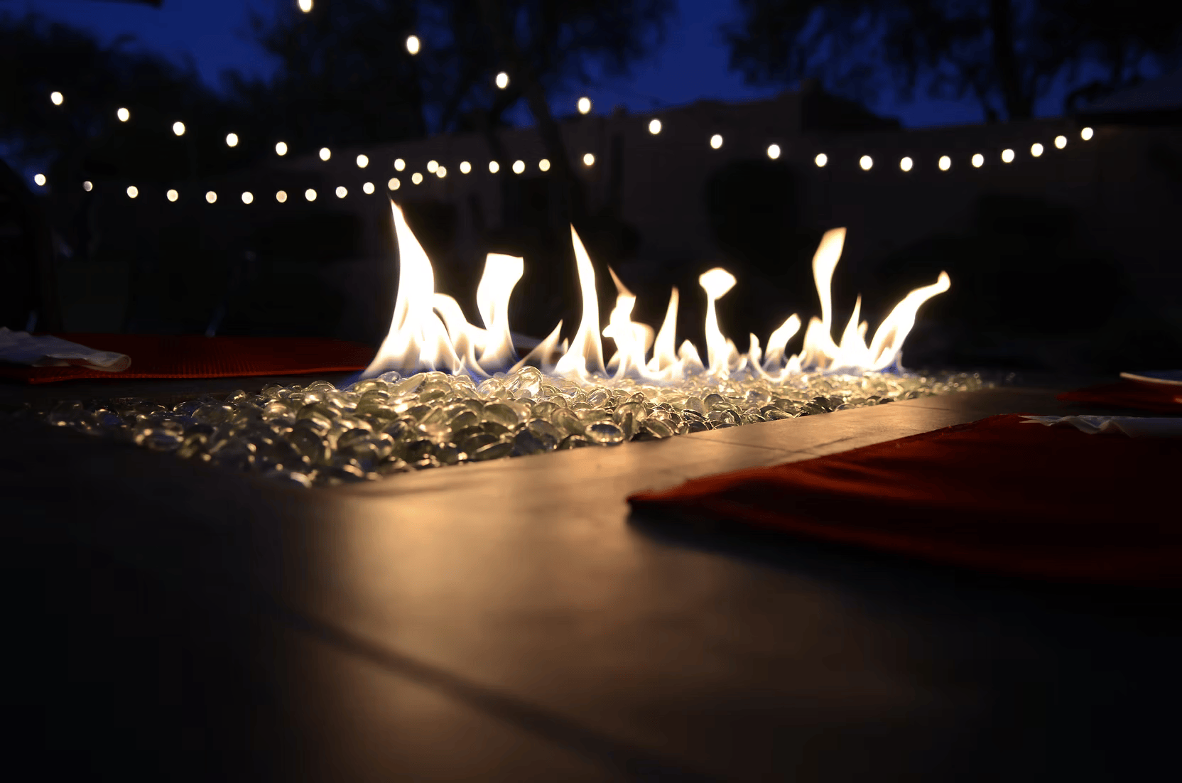 The Kokomo Grills Entertainer Bar Gas Fire Pit Table with Fire-Glass by Kokomo Grills glows with lively flames, surrounded by string lights at night, creating a warm, inviting outdoor scene beneath a dark blue sky and blurred trees.