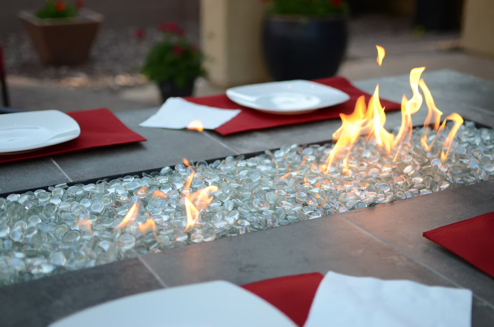 The Kokomo Grills Entertainer Bar Gas Fire Pit Table with Fire-Glass sits at the center of an outdoor dining table set with white plates, red placemats, and napkins, while potted plants blur in the background.