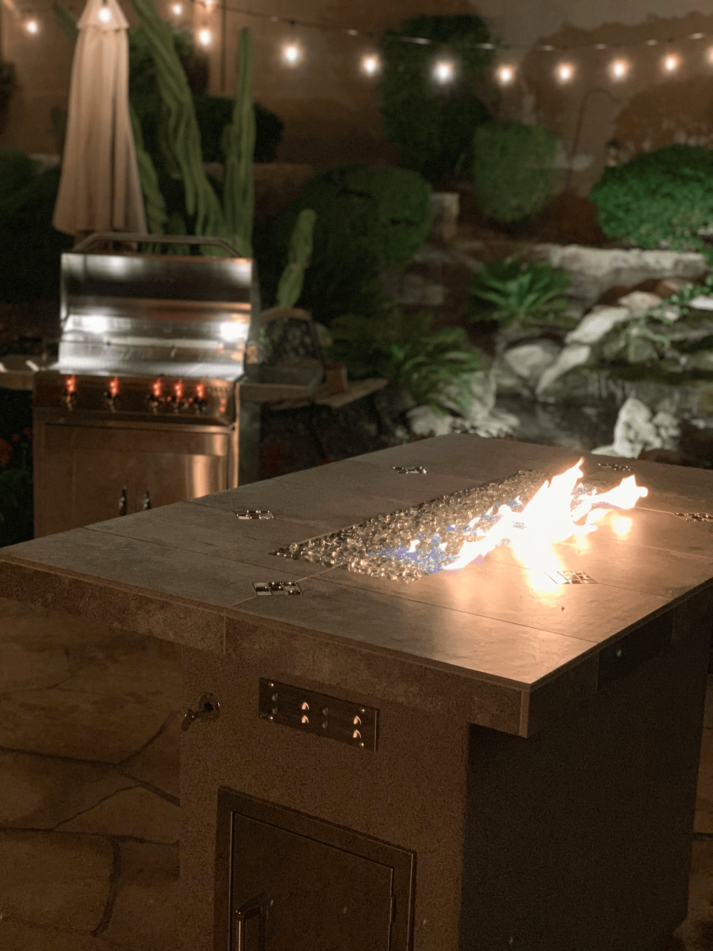 At night, a modern patio showcases the Kokomo Grills Entertainer Bar Gas Fire Pit Table with fire-glass in front and a Kokomo Grills stainless steel barbecue grill warmly lit in the background, all under glowing string lights overhead.