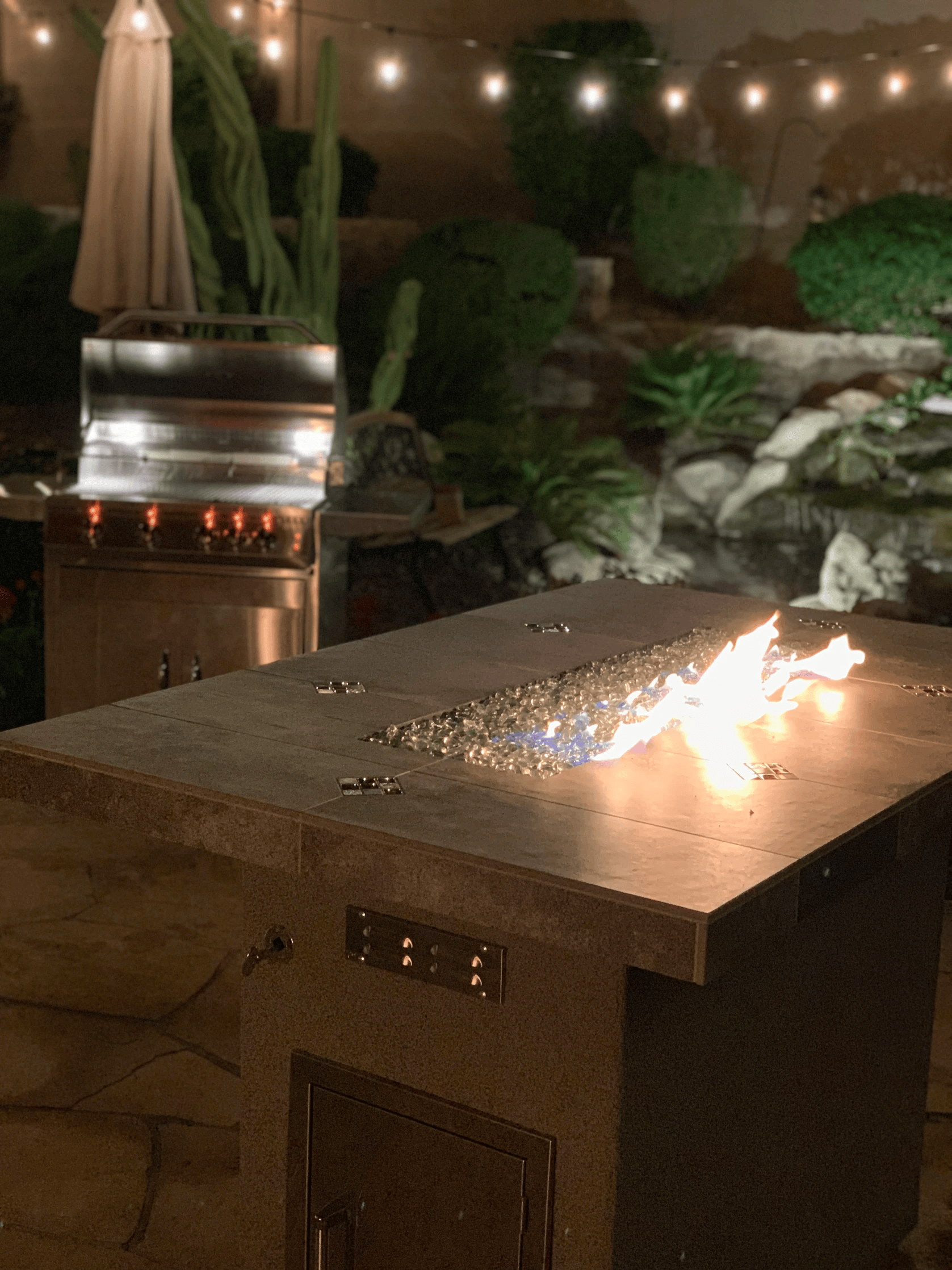 At night, a modern patio showcases the Kokomo Grills Entertainer Bar Gas Fire Pit Table with fire-glass in front and a Kokomo Grills stainless steel barbecue grill warmly lit in the background, all under glowing string lights overhead.