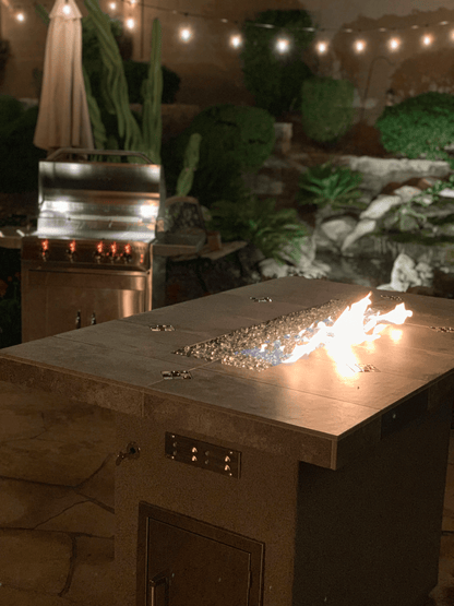 At night, a modern patio showcases the Kokomo Grills Entertainer Bar Gas Fire Pit Table with fire-glass in front and a Kokomo Grills stainless steel barbecue grill warmly lit in the background, all under glowing string lights overhead.