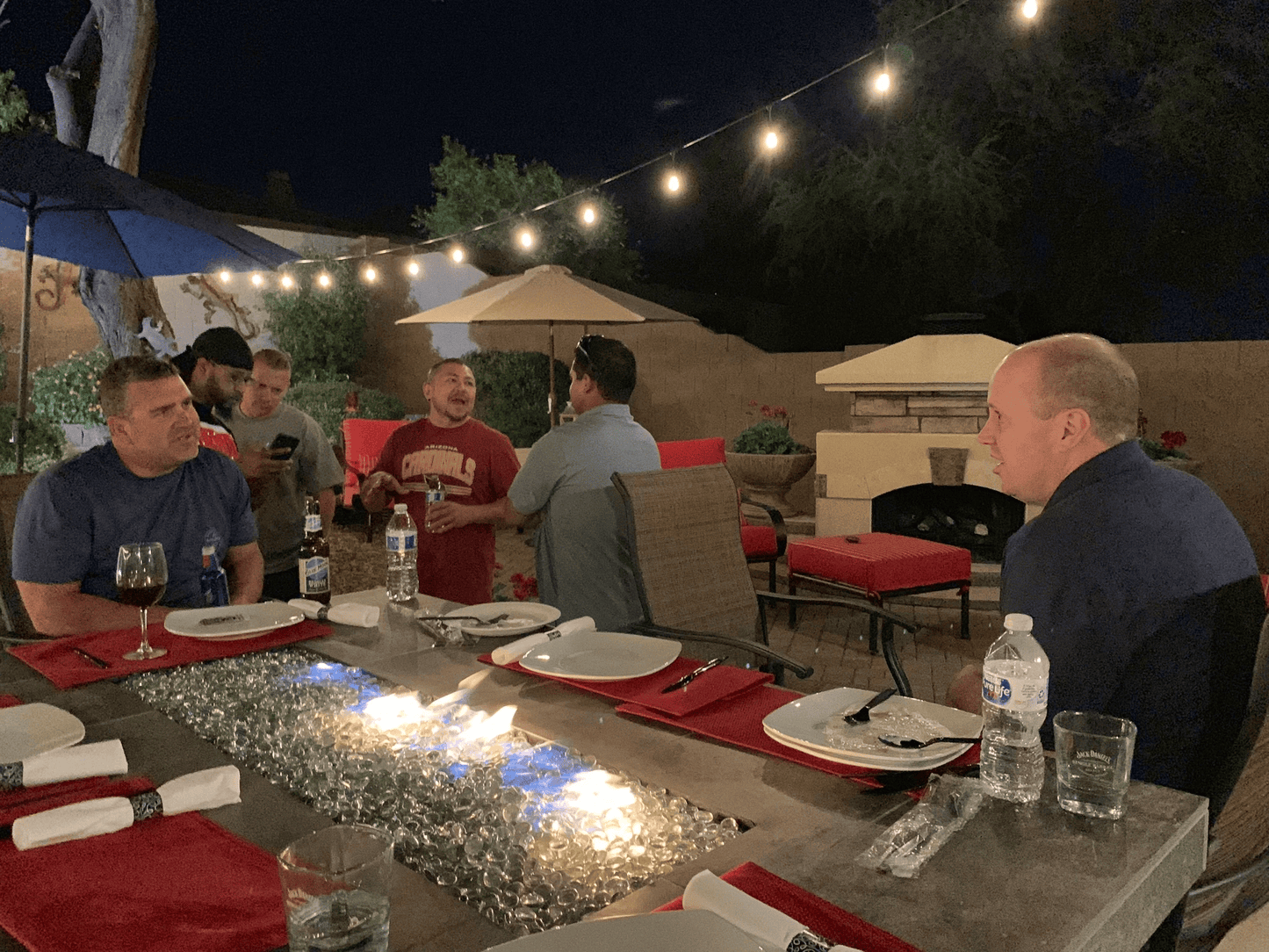 A group of men sit and chat around the Kokomo Grills Entertainer Bar Gas Fire Pit Table with Fire-Glass Entertainer-FP at night under string lights, with plates, napkins, drinks on the table, and a fireplace in the background.