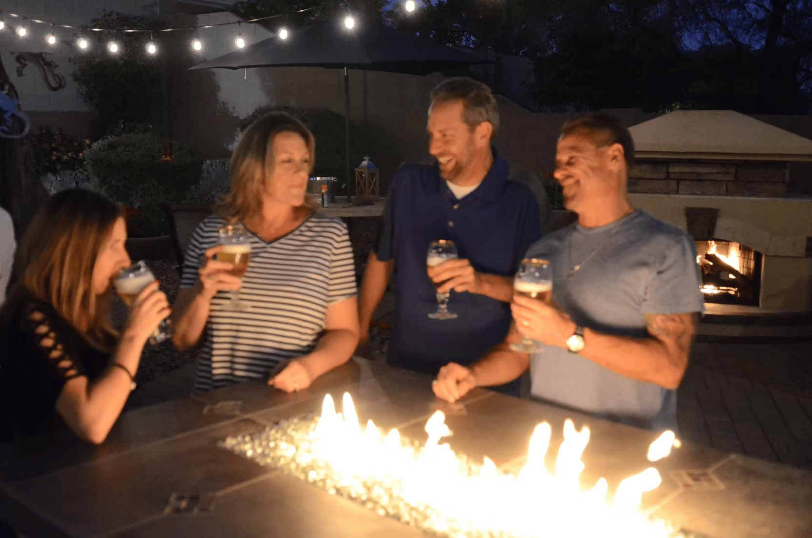 Four adults gather at night around the Kokomo Grills Entertainer Bar Gas Fire Pit Table with Fire-Glass, smiling and holding drinks. String lights and a pizza oven enhance the festive backyard atmosphere.