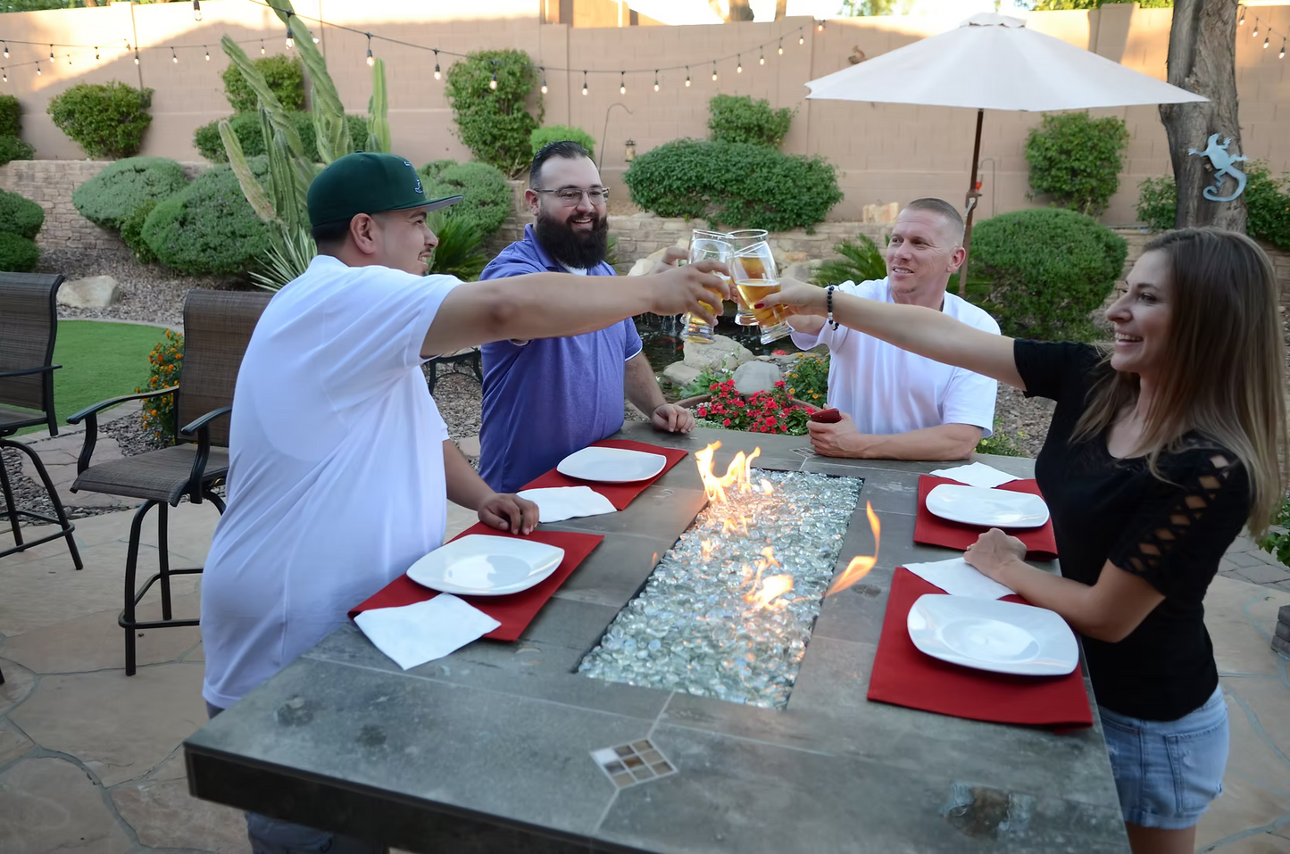 Four people gather around the Kokomo Grills Entertainer Bar Gas Fire Pit Table with Fire-Glass, raising glasses in a toast. Red placemats and empty plates sit on the table, while string lights and plants decorate the patio setting.