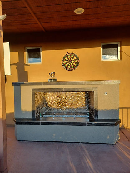 An outdoor patio showcases the Kokomo Grills Entertainer Fireplace LP or Nat Gas and Fire-Glass Burner Insert, with a mosaic tile interior, two small windows above, and a dartboard on the tan wall. Sunlight casts warm shadows across this cozy setting.