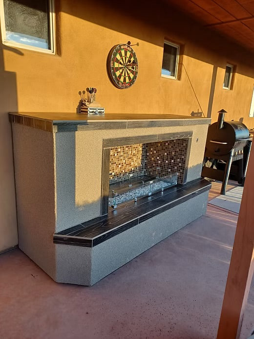 A Kokomo Grills Entertainer Fireplace LP or Nat Gas with Fire-Glass Burner Insert, featuring a mosaic tile inset and black stone trim, is set beneath a dartboard on a tan stucco wall beside a black grill on the covered patio.