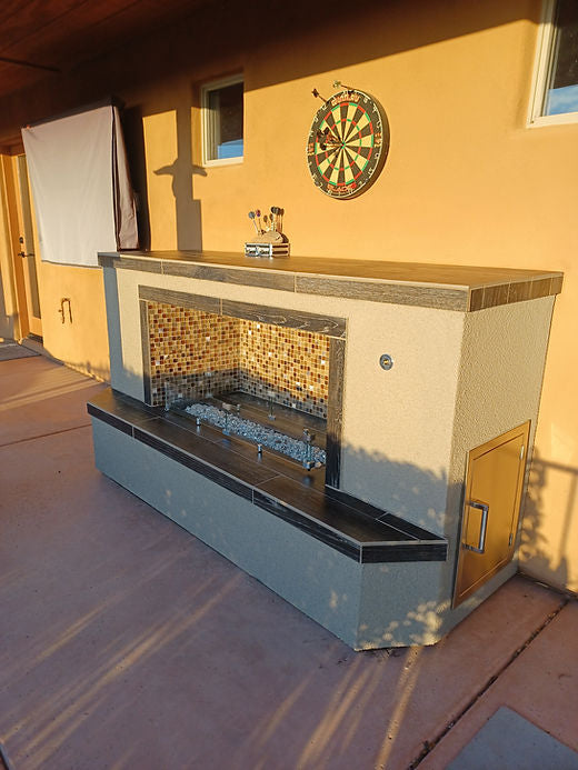 The Kokomo Grills Entertainer Fireplace LP or Nat Gas with Fire-Glass Burner Insert from Kokomo Grills is set against a stucco wall, topped by a dartboard and shelf for darts while sunlight stretches shadows across the patio floor.